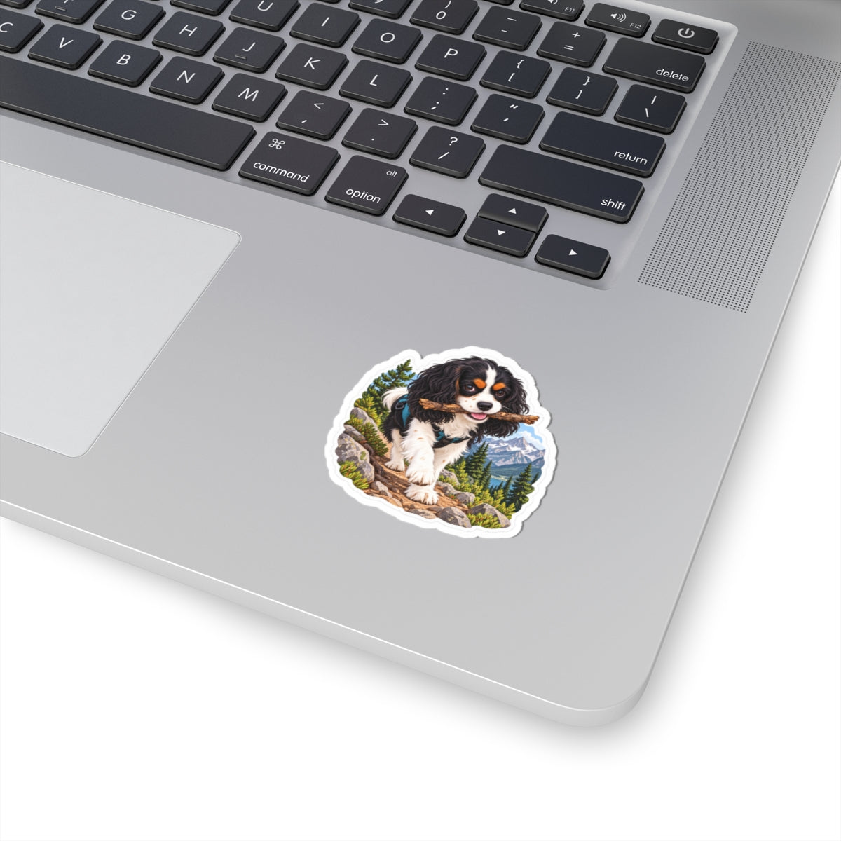 Product mockup of the Cavy Play Mode Trail Stick kiss-cut sticker: a tricolor Cavalier King Charles Spaniel carrying a stick along a mountain trail with pines and snowy peaks.